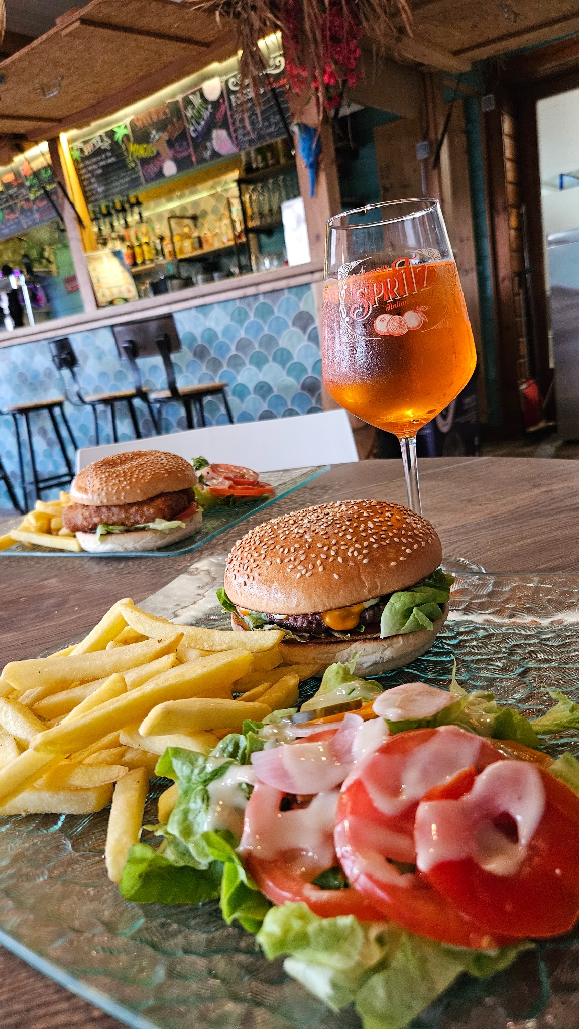 Burgers, frites et salade servis à table, avec un verre en arrière-plan au bar du Camping Ohana