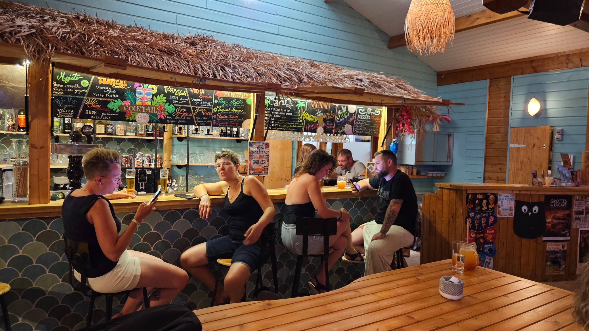 Des vacanciers réunis au comptoir du bar du Camping Ohana dans le décor en bois et turquoise