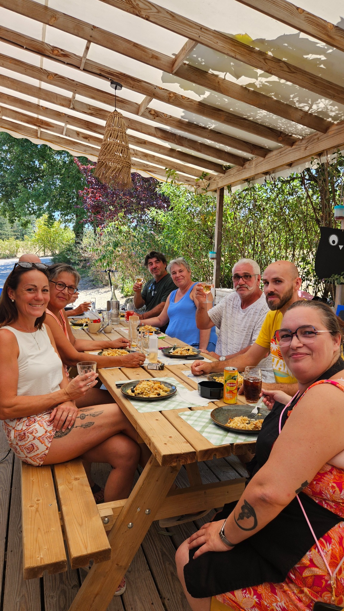 Un groupe de vacanciers partage un repas autour d’une grande table sur la terrasse couverte du Camping Ohana